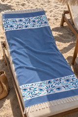 A blue beach towel with white and light blue floral and eye patterns and cream fringe, laid out on a wooden beach chair on sandy ground.